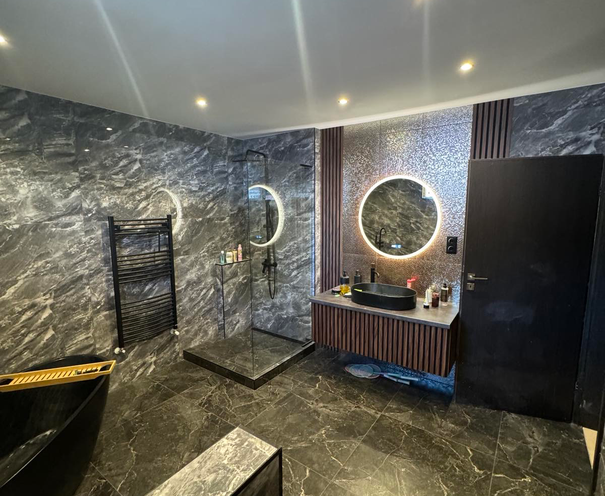 Bathroom tiled in black marble effect, featuring a circular mirror as the centerpiece, illuminated all around by a light.